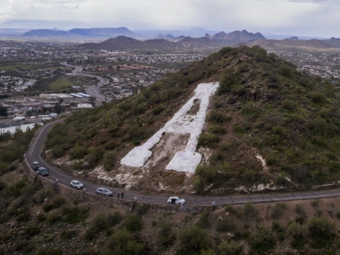 A scenic view of a mountain with a large white-painted 'A' on its slope, commonly known as 'A' Mountain. A winding road circles the base of the hill, with cars visible along the road. The backdrop includes a sprawling cityscape, surrounded by mountain ranges under a cloudy sky, with patches of blue visible.