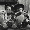 Two boys in cowboy hats sit side by side with their feet stretched out toward the camera, each reading a comic book. One holds The Ghost Rider while the other reads a Zane Grey western. The image is in black and white, highlighting their worn shoes and absorbed expressions.