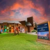 The exterior of the Flandrau Science Center and Planetarium at sunset. The building is constructed with red brick and features modern architectural designs, including a large dome that hints at its planetarium function. A series of rectangular windows are illuminated with various colors, creating a vibrant spectrum on the building's facade.