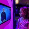 A young girl with long brown hair and a pink mermaid shirt gazes at an interactive screen in a science exhibit. The screen displays a man speaking against a blue molecular-patterned background. The exhibit is illuminated with vibrant purple and blue lighting.